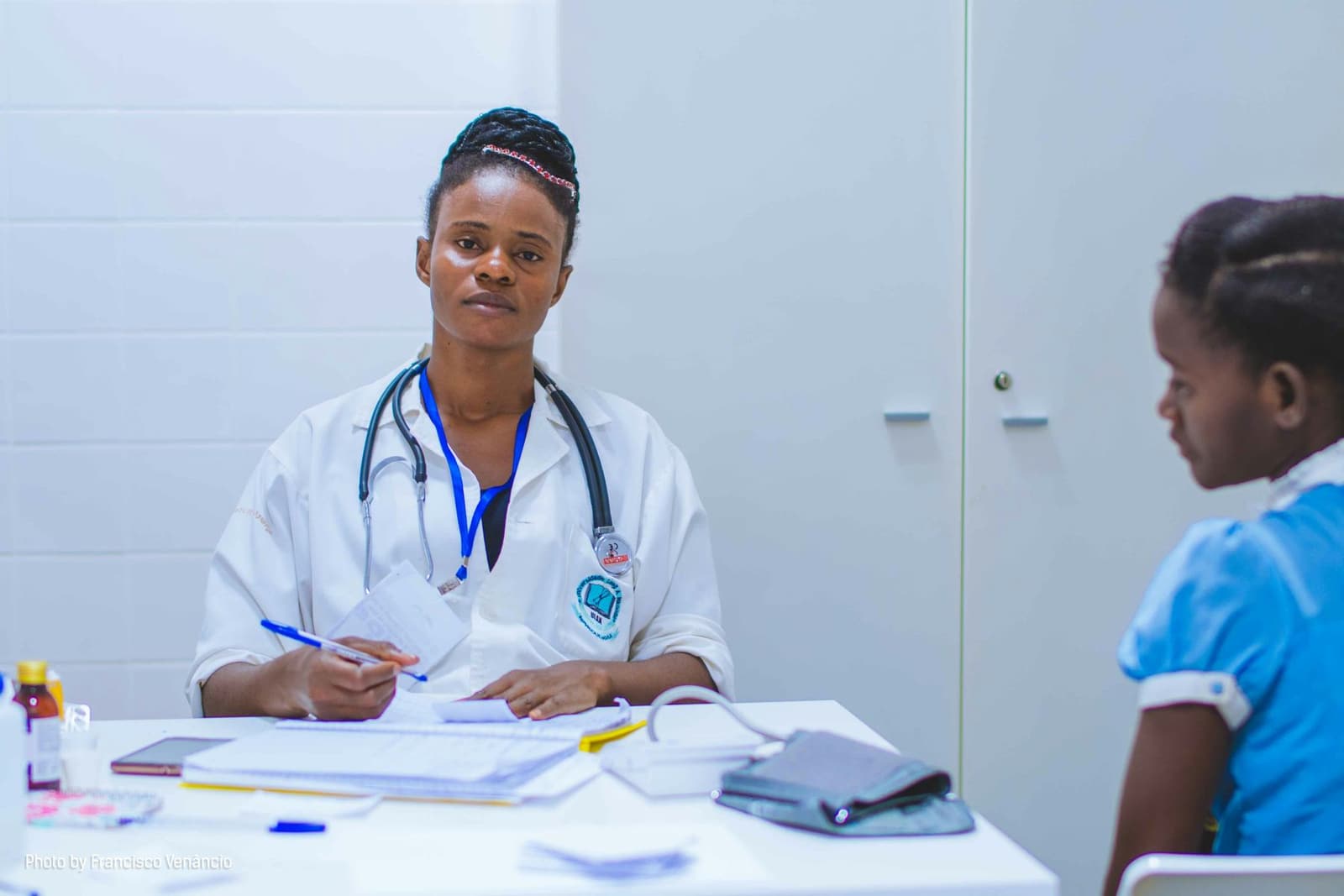 A Black female doctor in a white coat with a stethoscope talking with a young child at a desk