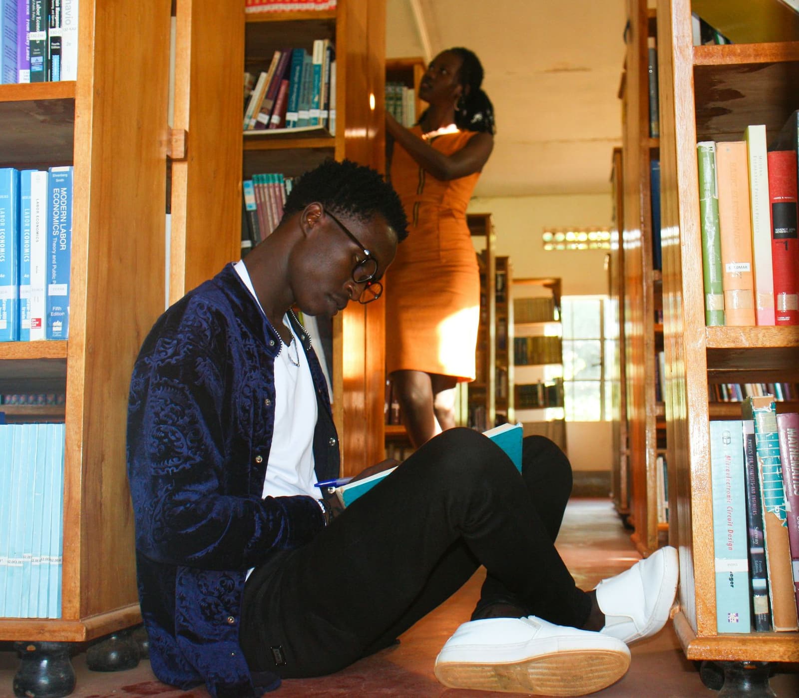 A young Black man sitting and reading in a library surrounded by bookshelves