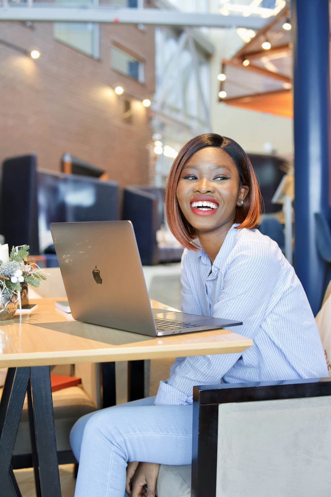 A smiling Black woman in a light shirt working on a laptop in a bright café