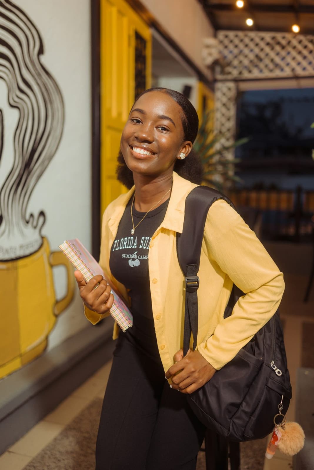 A smiling young Black woman student with a backpack, holding a notebook